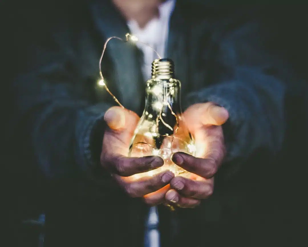 A person holds a glowing light bulb with small string lights inside, surrounded by darkness.