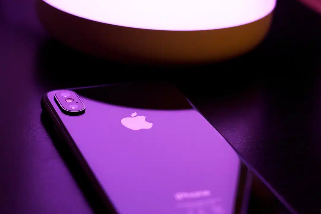 A black iPhone with a dual camera lies on a dark surface under purple lighting near a round lamp.