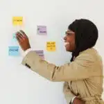 Smiling woman in a hijab placing sticky notes labeled "Zoom" and "Online" on a white wall.