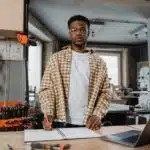 A man in a workshop stands at a workbench with blueprints and a laptop, wearing safety glasses and a plaid shirt.