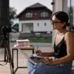 Woman with sunglasses sits indoors using a laptop, writing notes, and filming herself on a camera tripod.