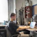 Man working on a laptop at a cluttered workbench in a workshop with tools and equipment around him.