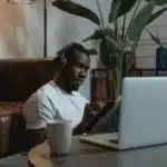 Man wearing headphones sits on floor by a sofa, writing in a notebook near a laptop and coffee mug.