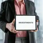 A person in a suit holds a tablet displaying the word "INVESTMENTS" on a white screen.