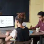 Three people work at a table, viewing a computer screen displaying a presentation with charts and graphs.