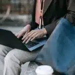 Person in business attire typing on a laptop outdoors, with a bag and a takeaway coffee cup nearby.