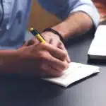 Person writing notes on paper beside a laptop on a desk.