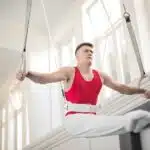 A male gymnast in a red tank top performs on still rings in a gym with white walls and large windows.