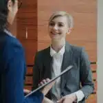 Two women in business attire talking; one is holding a clipboard, the other is smiling.