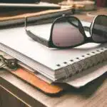 Black sunglasses resting on a spiral notebook and clipboard on a wooden desk.
