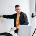 Man teaching math in a classroom, pointing at equations on a whiteboard during an online lesson.