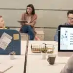 Four people collaborate in a modern office with computers, notes, and a presentation on charts and graphs.