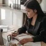 Woman in a black blazer works at a desk with a laptop and open notebook, appearing focused.