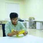Man in glasses reading a yellow book at a white table in a modern office with empty desks.