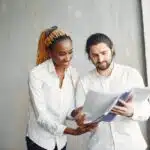 Two coworkers in white shirts smiling and looking at documents together in a modern office setting.