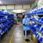 A woman in blue uniform organizes items in blue bins on shelves inside a warehouse or storage facility.