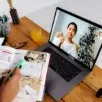 Person studies with a textbook while video chatting with a teacher giving a thumbs up on a laptop.