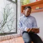 Man in a blue shirt sits by a window, reading a tablet in a bright, modern room with shelves and a plant.