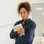 Woman in striped shirt holding books, standing in a bright hallway with a confident expression.
