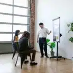 Person giving a presentation on a whiteboard to two seated people in a modern, bright room with plants.