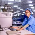 Smiling woman in blue uniform works at a computer in a brightly lit industrial or factory setting.