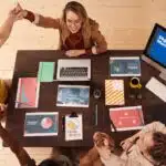 Four people celebrate around a table with laptops, papers, and a screen displaying "Digital Marketing.
