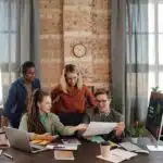 Four people gathered around a desk in an office, reviewing documents and discussing work together.