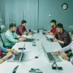 Six people working on laptops around a conference table in a modern office meeting room.