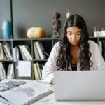 Woman with long dark hair studying at a desk with a laptop, books, and notebooks in front of a bookshelf.
