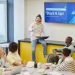 Man presenting "Start It Up!" slideshow to four colleagues in a modern office meeting room.