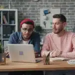 Two men working together at a desk with a laptop, notebooks, and color swatches in a modern office.