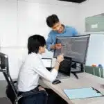 Two people discussing code shown on a computer monitor in an office setting with desks and chairs.