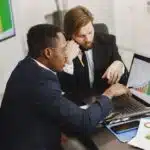 Two businessmen in suits analyzing colorful bar graphs on a laptop at an office table with documents.