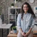 Smiling woman with long curly hair stands in a modern office by a desk with a laptop and plants.