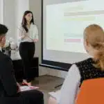 Woman presenting a bar graph on a projector screen to two seated colleagues in a meeting room.