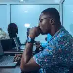 Man in floral shirt works thoughtfully on a laptop in an office with others at computers.