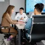Four people sit around a conference table having a discussion in a modern office with a digital screen.