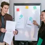 Two people smiling and sharing charts and sketches in front of a whiteboard with colorful sticky notes.