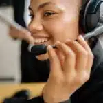 Smiling woman wearing a headset with microphone, holding the mic, and looking ahead, possibly at a screen.