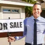 Man in dress shirt and tie holding a "FOR SALE" sign in front of a house with a garage.