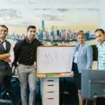 Four people stand in an office smiling next to a whiteboard that reads "NEW HIRE!!!" in red letters.