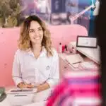 Smiling woman in white shirt sitting at a desk, talking to someone across from her in an office setting.