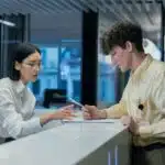 Two people at a modern reception desk discussing documents, one holding a pen and paper, the other gesturing.