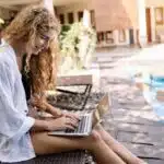 Woman sitting poolside outdoors, smiling while typing on a laptop, wearing a white shirt and shorts.