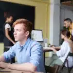 Young man working on a laptop in an office with three colleagues collaborating in the background.