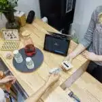 Customer pays with a card at a wooden counter while a cashier holds a card reader; drinks are on the counter.