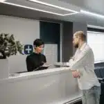 A man speaks to a receptionist at a modern clinic reception desk with chairs and a potted plant nearby.