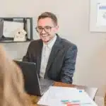 Smiling man in a suit sits at a desk with a laptop, talking to someone across from him.