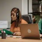 Two women wearing headsets work at laptops in a call center office, with folders and stationery on the desk.