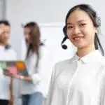 Woman in a headset smiling at the camera, with two colleagues talking in the blurred background.